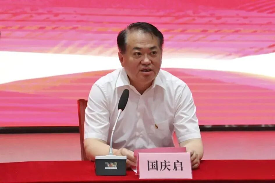 一位男性在会议上发言，穿着白色衬衫，桌上有一个麦克风和他的名字牌，背景为红色。
