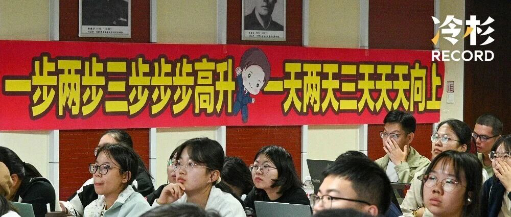 一组学生在听演讲，背景有一条红色横幅，上面写有激励语句，旁边有卡通角色