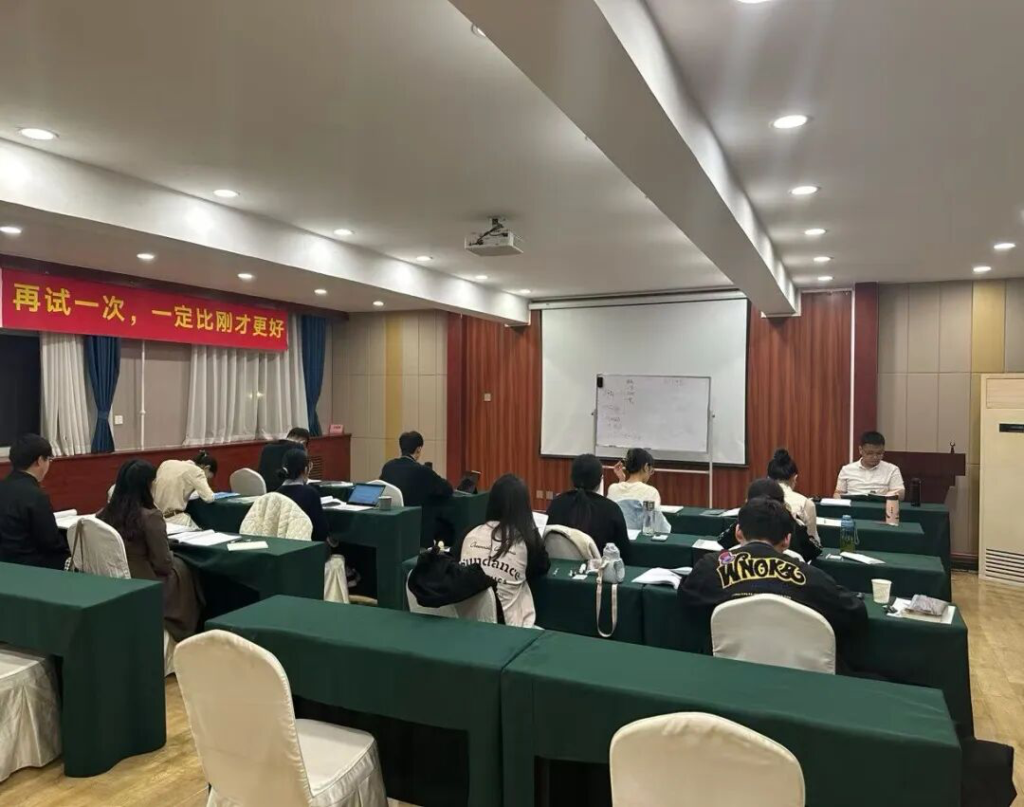 一个课堂场景，几位学生坐在绿色桌子前，专注听讲，前方有投影仪和白板，墙上挂着红色标语。
