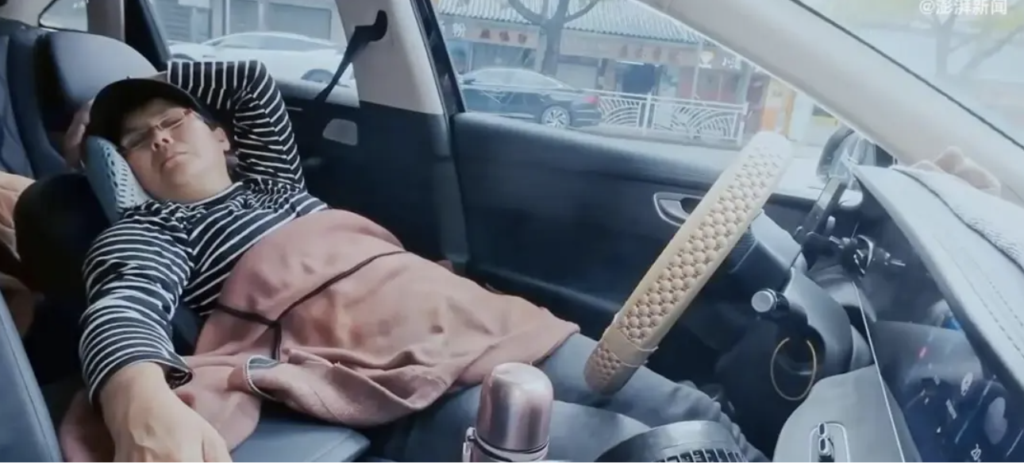 A man sleeping in a car, wearing a striped shirt and glasses, with his arms resting behind his head. The interior of the car is visible, showing a steering wheel and dashboard.